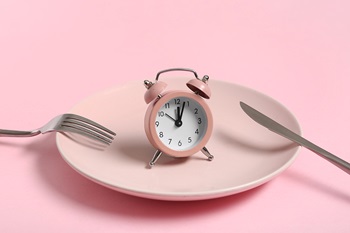 Clock plate Light pink plate with a clock in the middle and a fork and knife on a pink background.