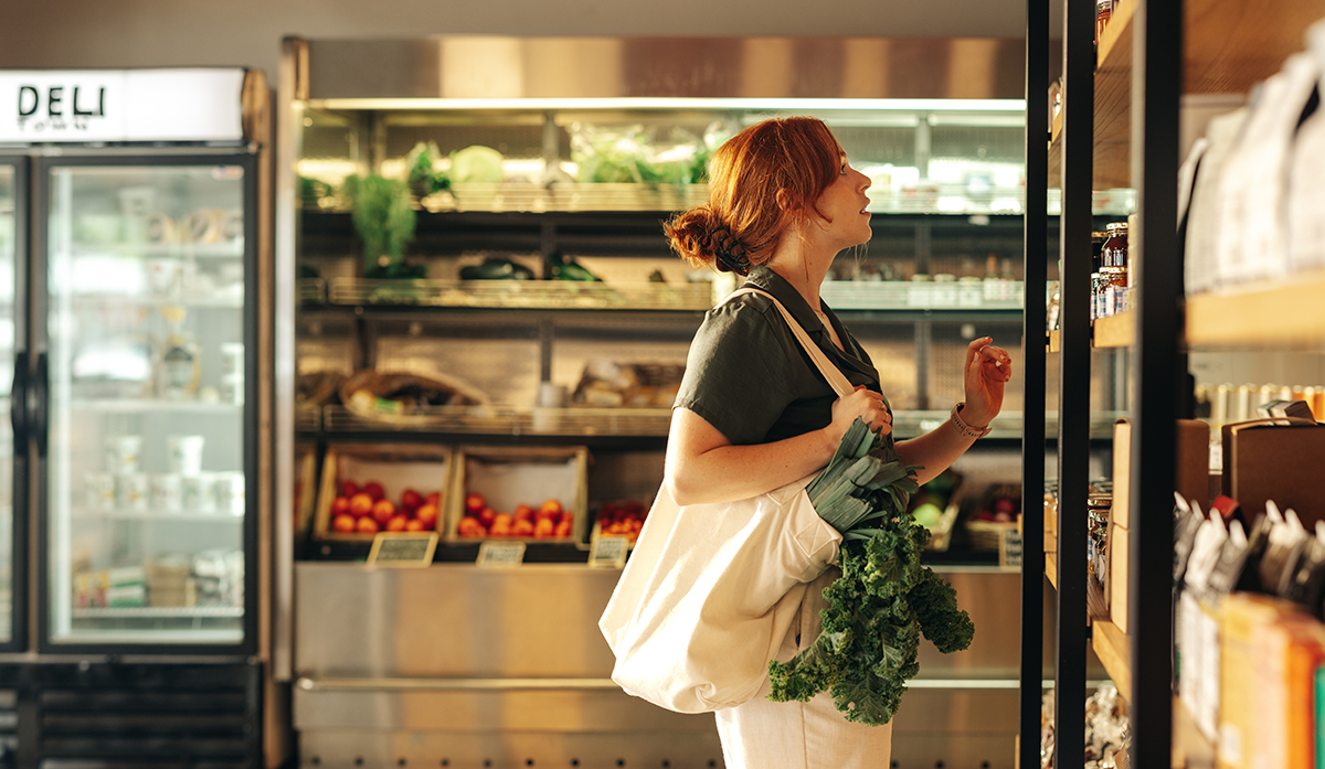 Woman in grocery store holding a reusable bag with produce in it looking at products on a shelf and trying to decide what to get